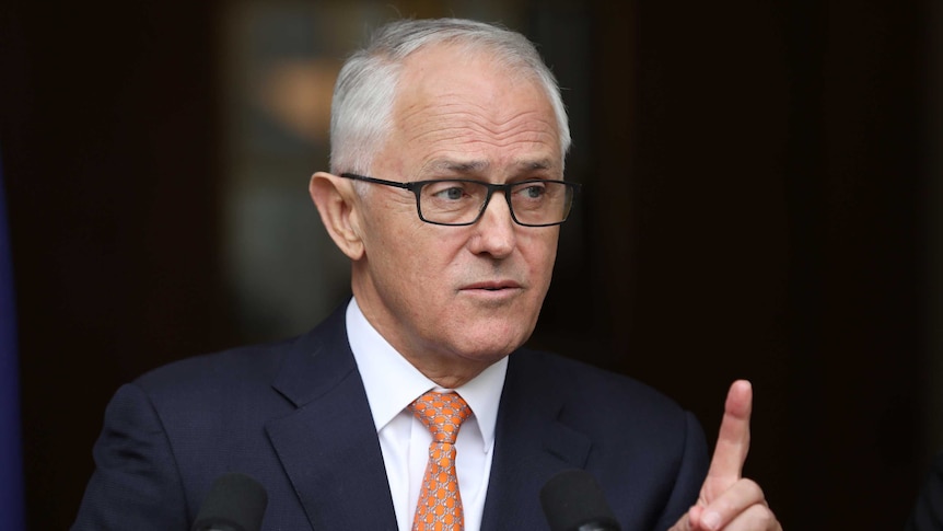 Turnbull in front of a dark background with one finger raised looking left to right, wearing a navy blue suit and orange tie.