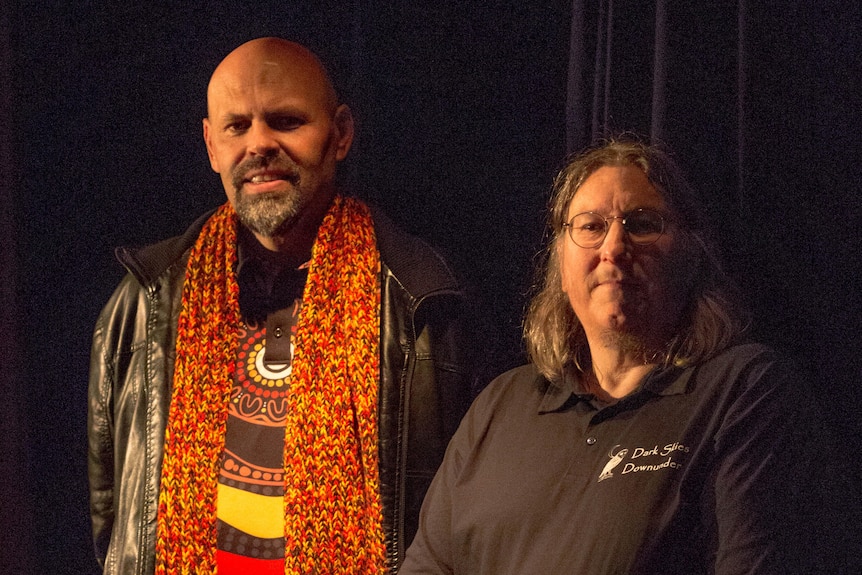 Two men, one draped in a bright orange scarf and T shirt with Inidgenous artwork, stand in a dark theatre.