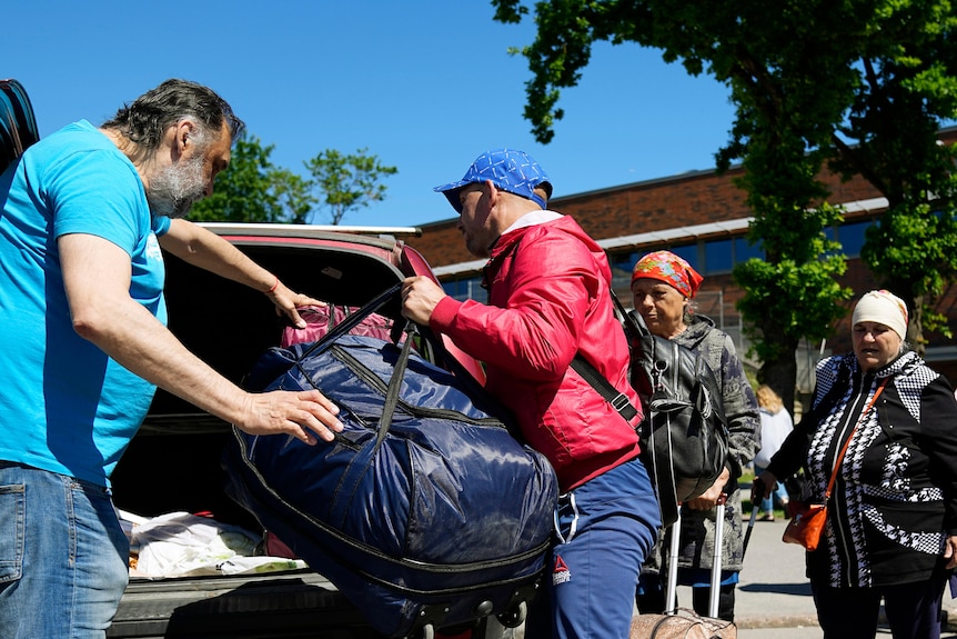 A man in a red jacket loads luggage into the back of a car as a Ukrainian family watches on.