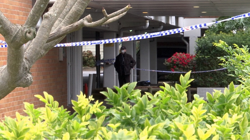 A police officer in a mask stands at the front door of a brick building that's been cordoned off with tape.