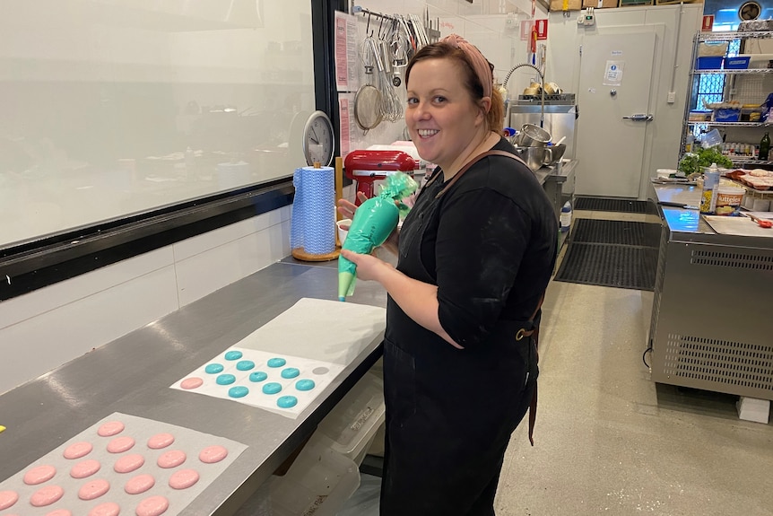 A woman is seen looking to the camera and smiling as she pipes macaroon shells in a commercial kitchen. She wears black.