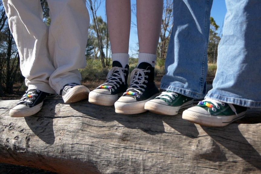 Three people's shoes, standing on a log, all with rainbow beads threaded on their laces.