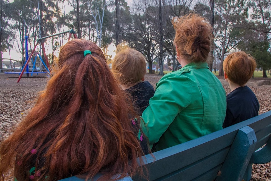 Back of the head shot of three children and a woman, all with red hair, sitting on a park bench