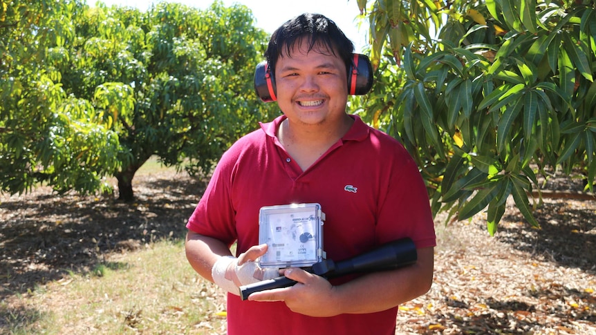 Han Shiong Siah holding a little goose-buster speaker and a laser torch in front of a mango orchard.