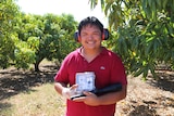 Han Shiong Siah holding a little goose-buster speaker and a laser torch in front of a mango orchard.