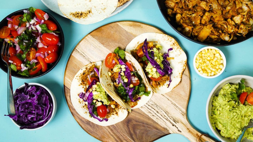 Three barbecue pulled jackfruit tacos on a platter surrounded by salsa, cabbage, guacamole, pulled jackfruit and corn tortillas.