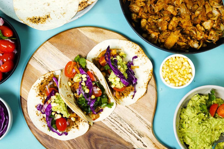 Three barbecue pulled jackfruit tacos on a platter surrounded by salsa, cabbage, guacamole, pulled jackfruit and corn tortillas.