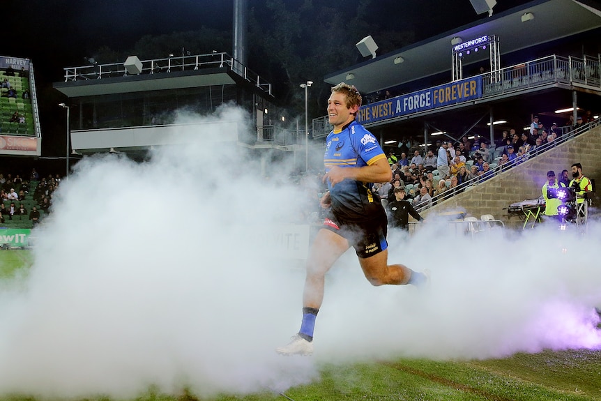 A Western Force rugby union player runs out onto the pitch, surrounded by dry ice with a home crowd in the stand behind.