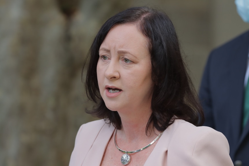 A woman with brown hair speaks while wearing a pale pink jacket and a large pendant necklace.