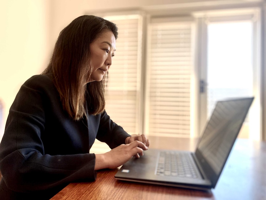 A woman of Asian decent works on her laptop. She has long hair and is wearing a black jacket.