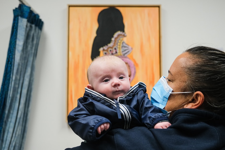 A midwife wearing a facemask holds a little baby in her arms.