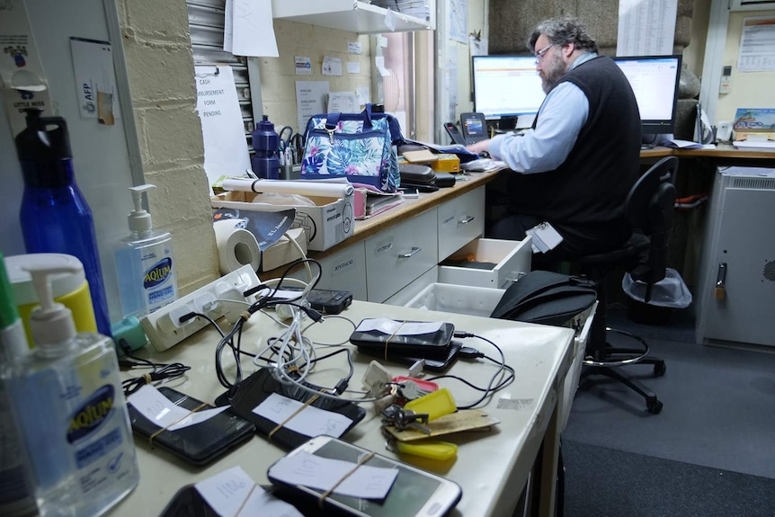 A large amount of phones sit on charge, while Brendan Podger works in the background.