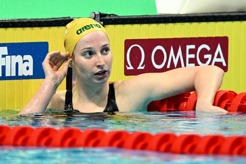 An Australian swimmer leans on the lane rope looking up at the screen with her mouth open. 