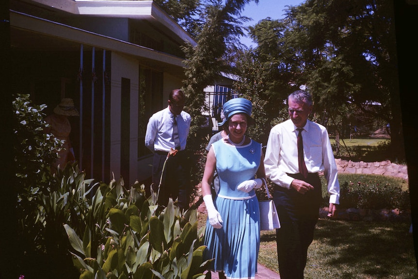 Vieille photo de la reine et du prince marchant bras dessus bras dessous dans un jardin avec un bâtiment en arrière-plan.