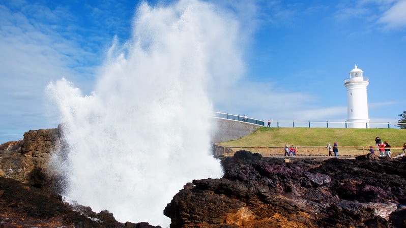 A spectacular blast of seafoam explodes out of a blowhole in front a lighthouse standing courageously on a headland.