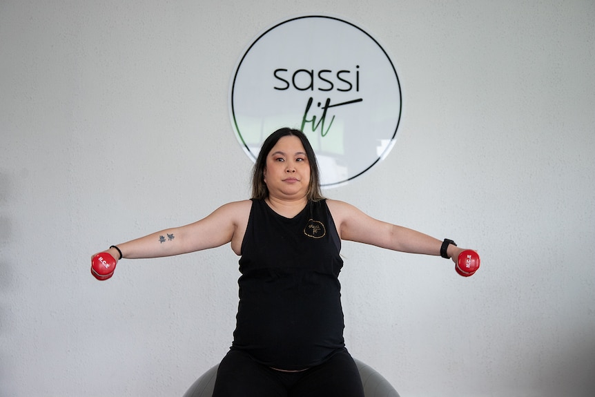 A pregnant Asian woman wearing a black singlet has her arms outstretched holding red hand weights