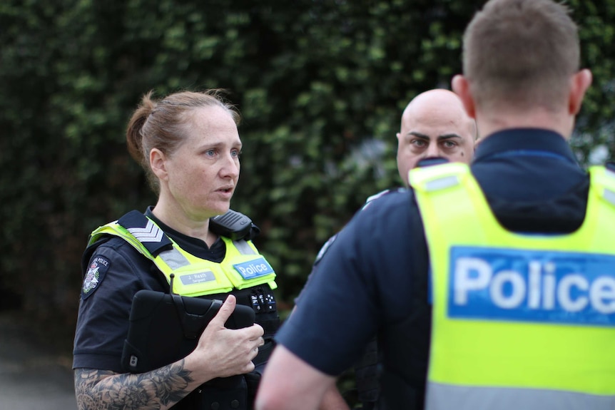 A female police officer holding an iPad looks towards a person who is obscured by her male colleague.