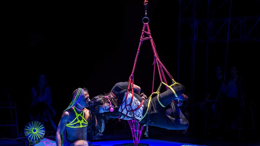 A man is tied up and hanging on stage while another man supervises
