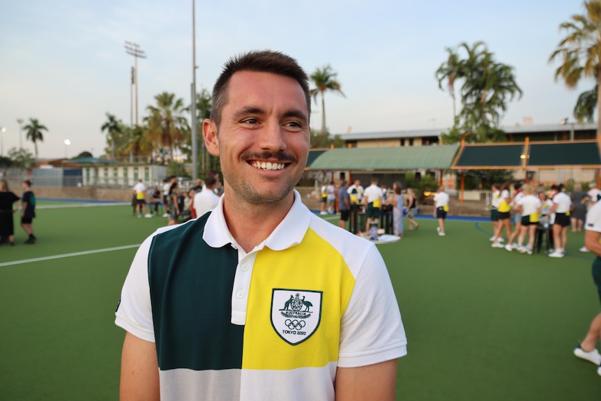 Jeremy Hayward wears a green and gold polo and smiles in a hockey field.