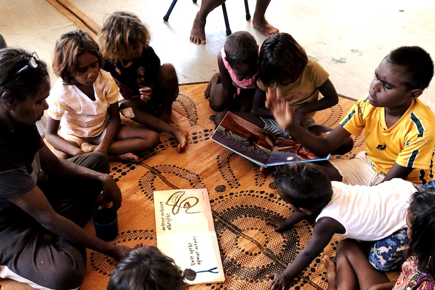 A woman and six children sit in a circle on a mat, reading picture books together.