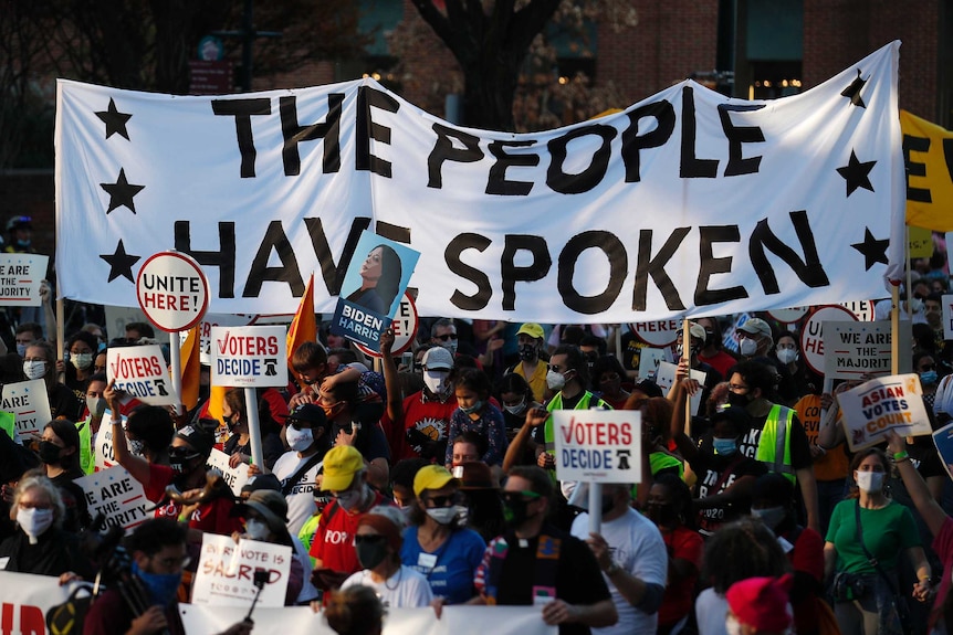 A crowd of people marching in the streets, holding signs saying "the people have spoken" and "voters decide".