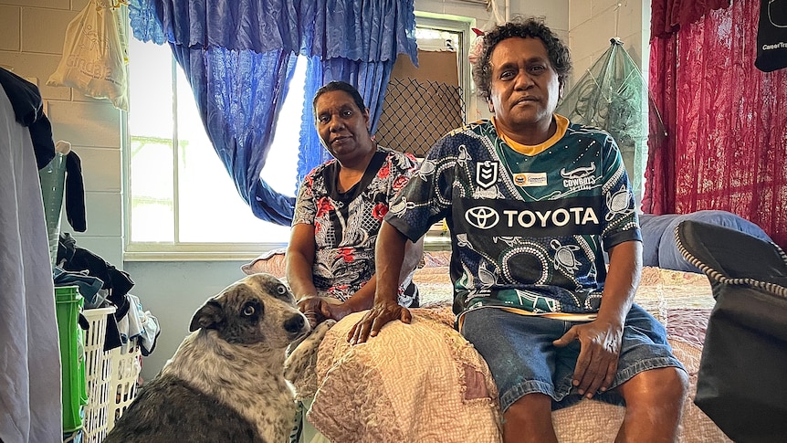 Denise and Colin Cedric sit on a bed, with their family dog, in a bedroom with colourful fabric drawn across the windows.