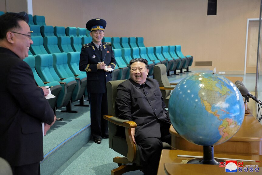 Kim Jong-un with a big smile on his face sits at a desk with a globe while flanked by two other officials