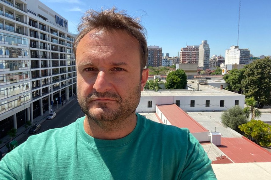 A close-up of a man standing outdoors with buildings in the background. 