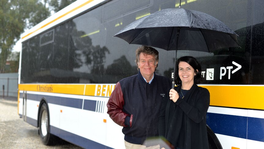 General Manager Glenn Christian and his daughter Rebecca Christian stand under an umbrella  in front of a stationary bus.