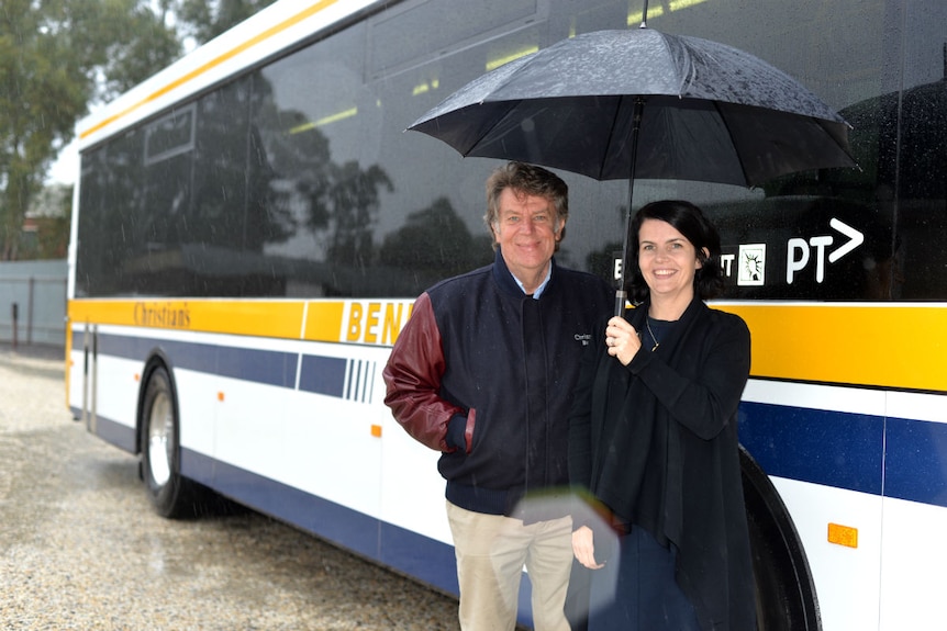 General Manager Glenn Christian and his daughter Rebecca Christian stand under an umbrella  in front of a stationary bus.