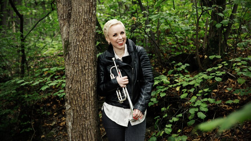 Trumpeter Tine Thing Helseth, wearing a black leather jacket, stands next to a tree in a forest holding her trumpet.