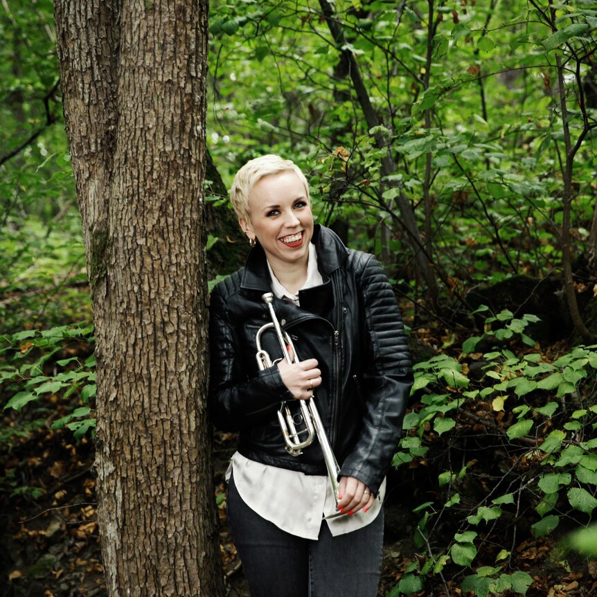 Trumpeter Tine Thing Helseth, wearing a black leather jacket, stands next to a tree in a forest holding her trumpet.