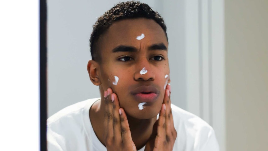 A young man with dark skin is looking in a mirror while putting moisturiser cream on his face with his hands.