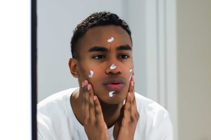 A young man with dark skin is looking in a mirror while putting moisturiser cream on his face with his hands.