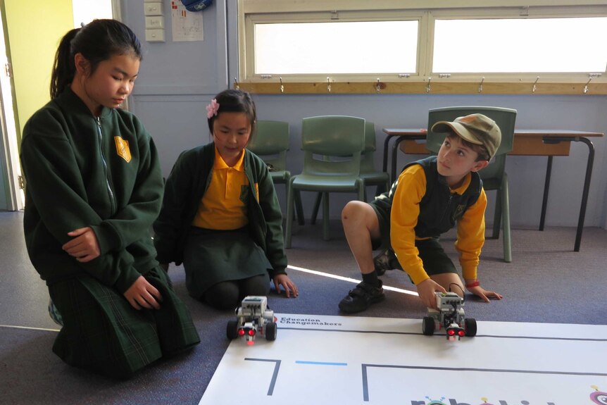 Three primary school students kneel on the ground in a classroom looking down at two small robots.