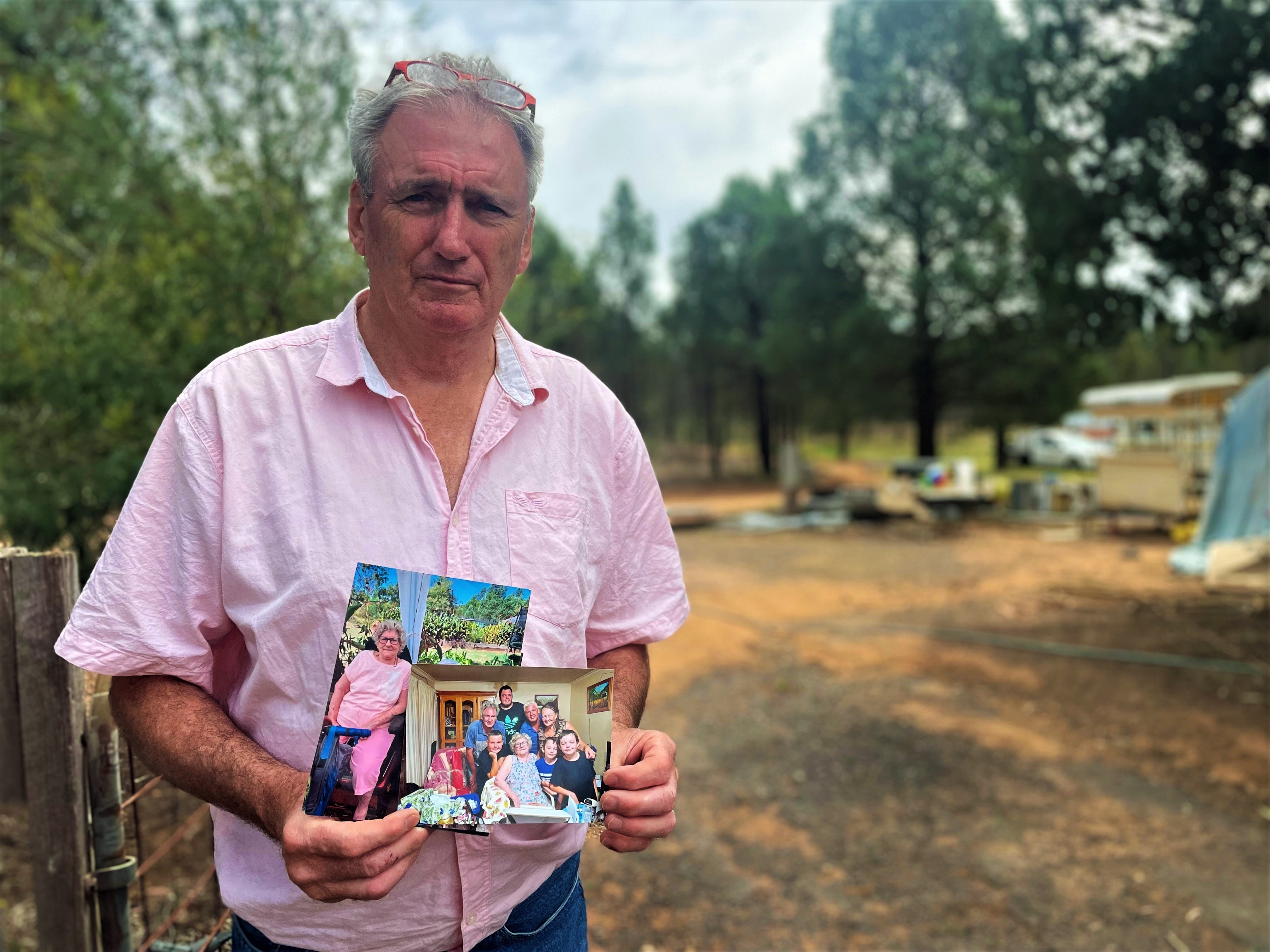 A man stands holding two pictures of his mother 