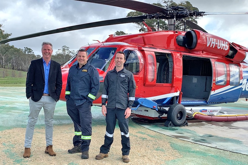 Three men stand in front of a red helicopter