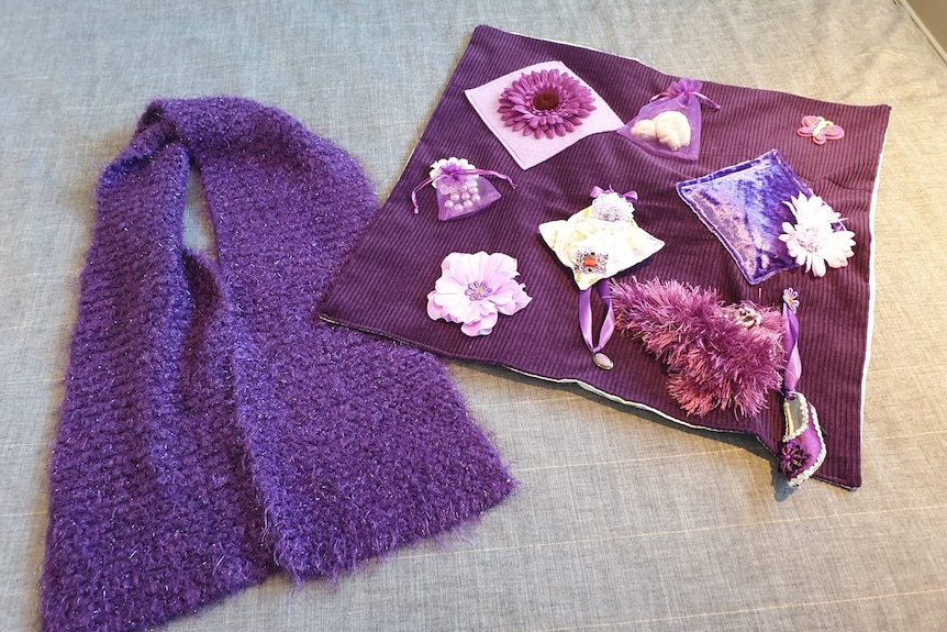 Purple woollen scarf laying beside a purple 'busy blanket' containing ribbons and flowers.