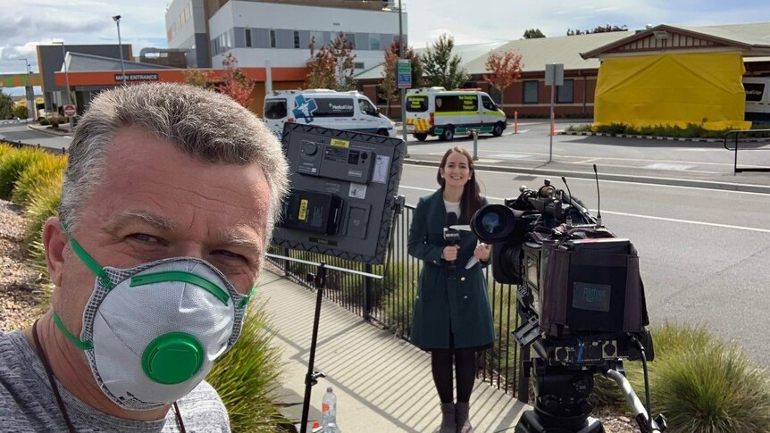 Cameraman wearing face mask and journalist standing in front of camera outside hospital.
