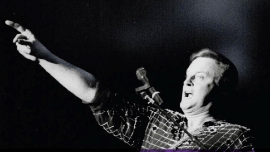 Singer, Peter Allen stands on a stage in front of a microphone with the words "The Peter Allen Festival " on the photo.