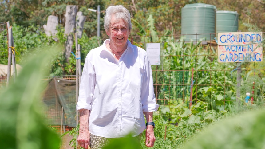 An smiling 82-year-old woman stands in a green garden. A sign behind her says 'Grounded Women Gardening'.