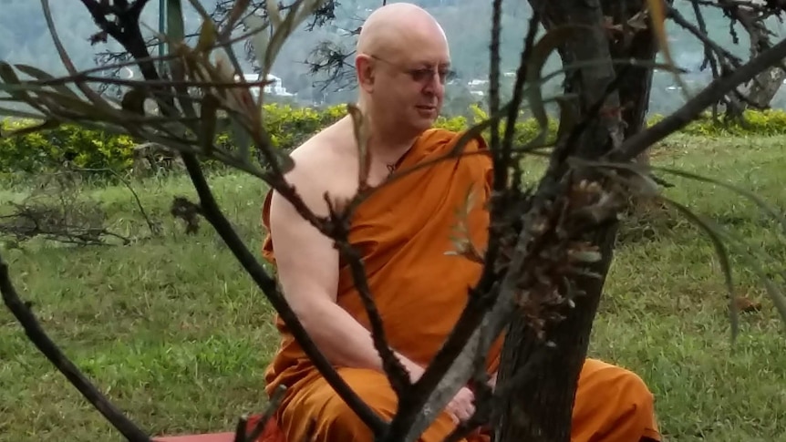 A Buddhist monk in saffron robes sits on the grass with tree branches in the foreground.