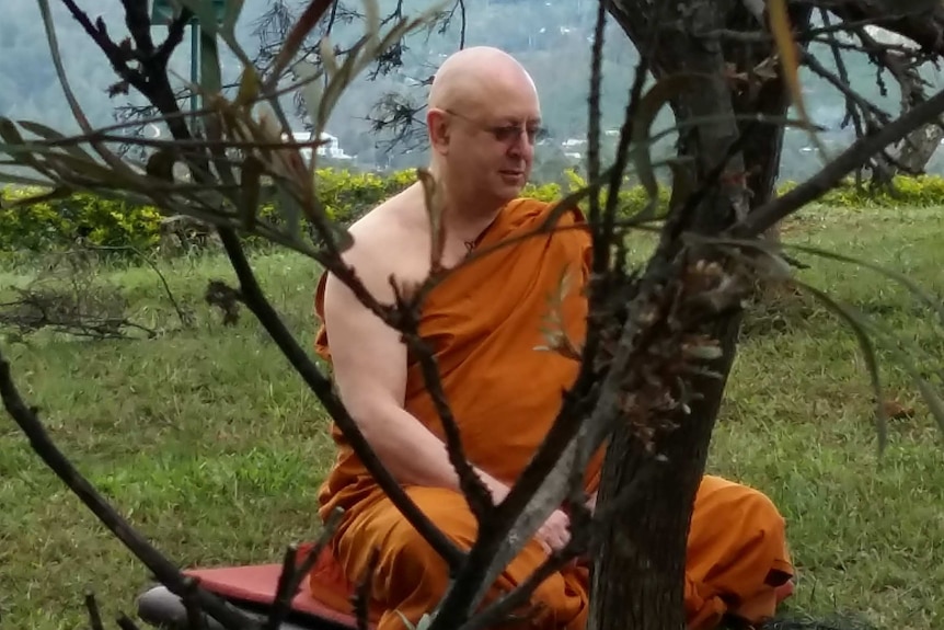 A Buddhist monk in saffron robes sits on the grass with tree branches in the foreground.