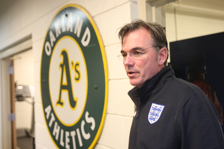 Billy Beane stands in the clubhouse next to an Oakland Athletics sign.