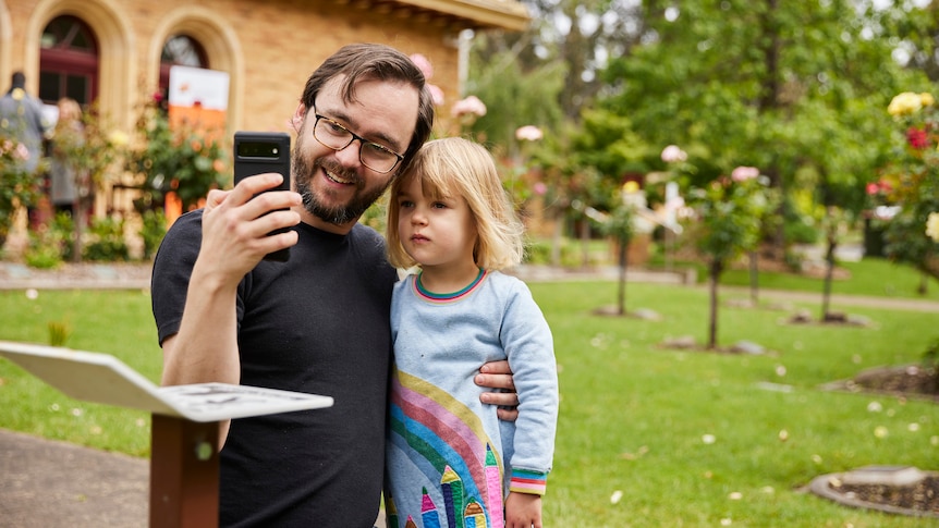 A smiling man wearing a black shirt, holds his young daughter while he also holds a smart phone they both look at.