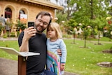 A smiling man wearing a black shirt, holds his young daughter while he also holds a smart phone they both look at.