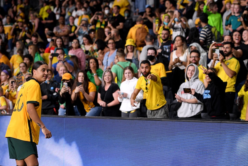 Sam Kerr of Matildas stands on edge of field and looks at fans over a blue scaffolding