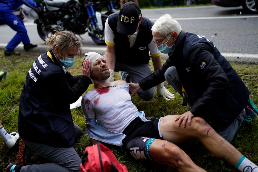 A cyclist with blood on his shirt and his head bandaged is looked after by medical staff on a roadside at the Tour de France.