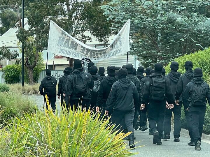 A group of men in black wearing balaclavas walk behind a banner.