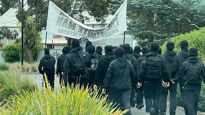 A group of men in black wearing balaclavas walk behind a banner.